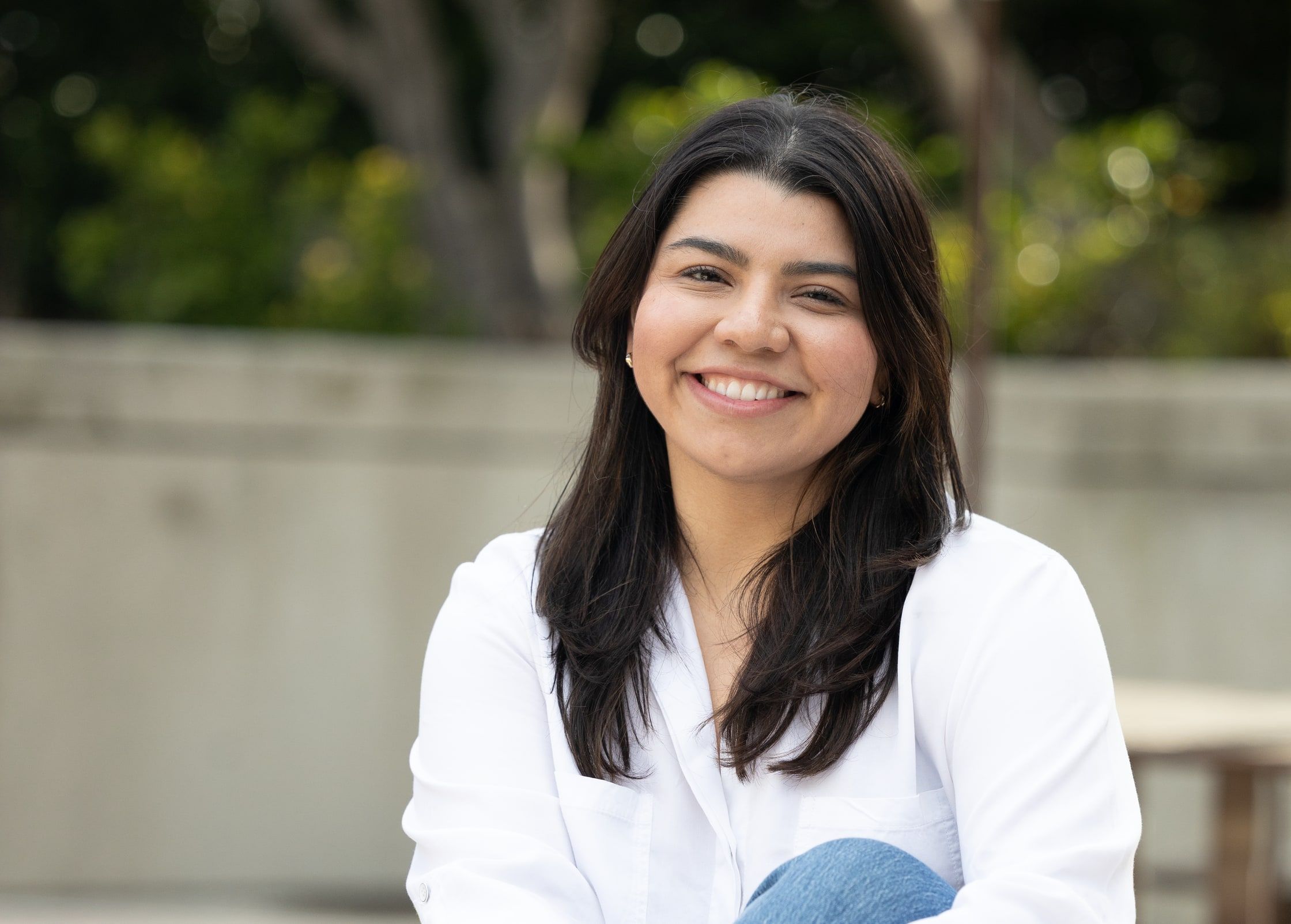 Kimberly Ramirez smiling, seated on a low wall with trees behind her